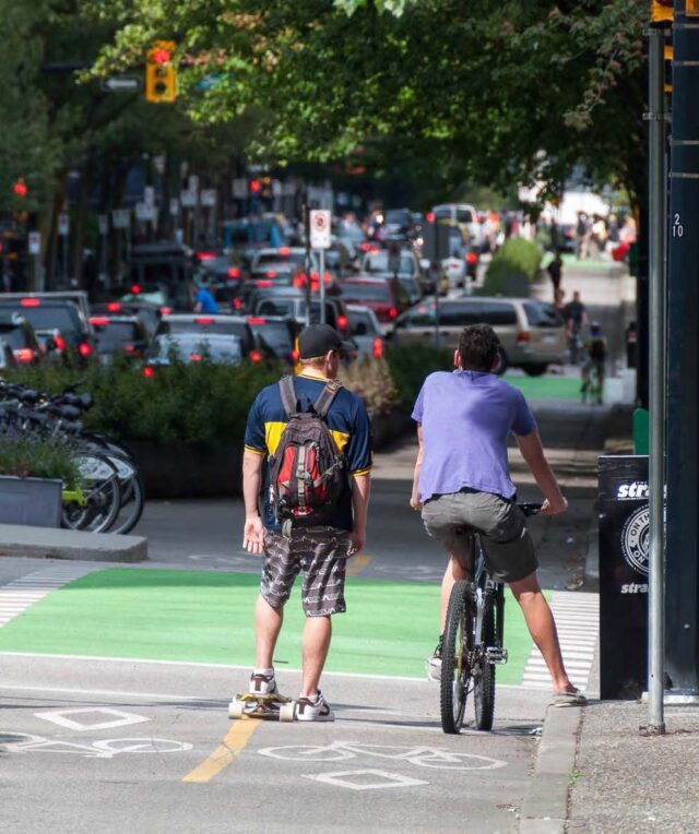 A longboard rider and a bike rider face away from the camera, waiting at a light to cross the street. They are riding in a two-way protected bike lane.