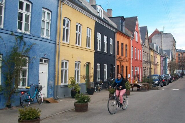 Claudia rides a bike on a quiet residential street. She wears pink pants, a black sweater, and a blue scarf.