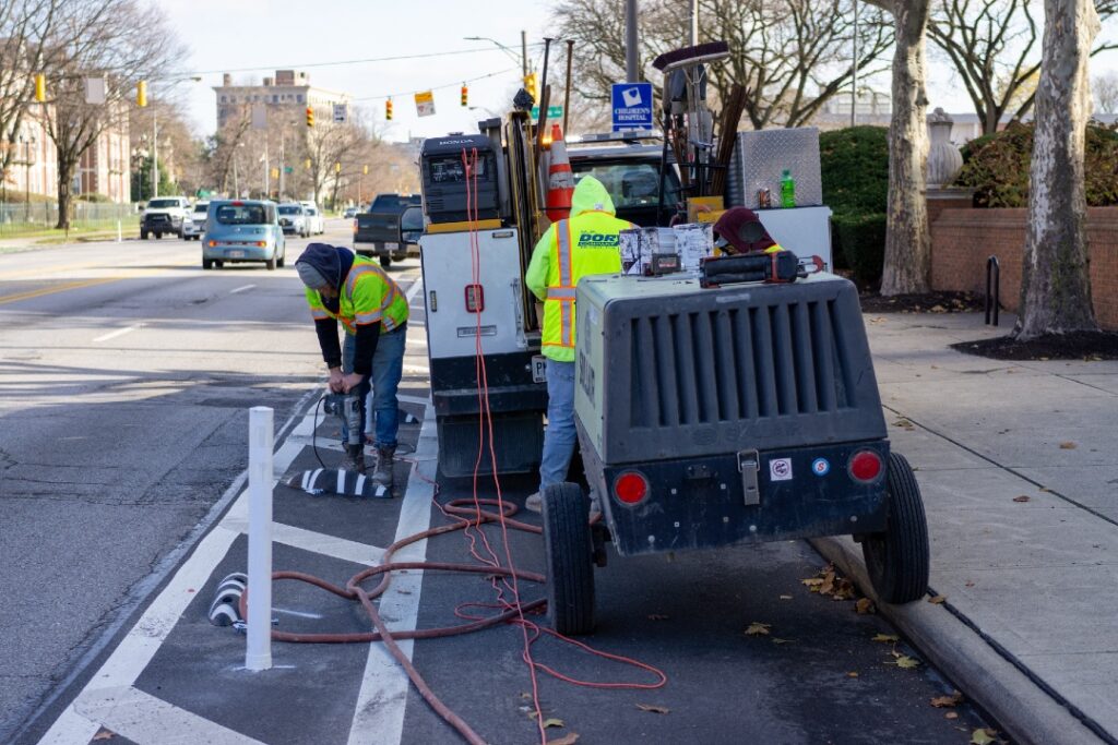 Three people in construction vests install a separated bike lane in Columbus Ohio. They are installing zebra-style bike lane separators next to a taller flex post.