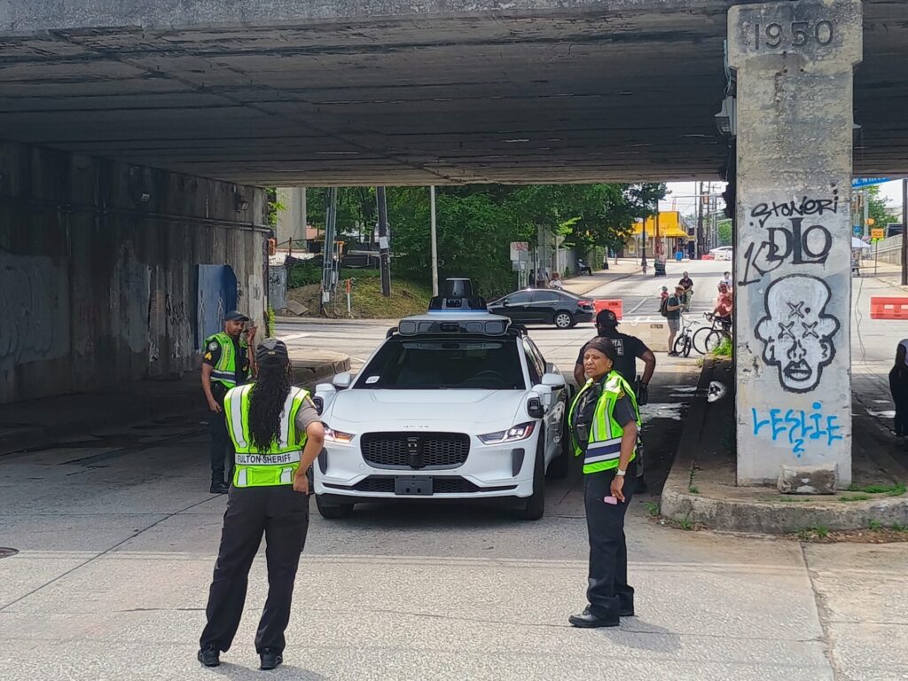 A Waymo vehicle enters an open streets event where vehicles are restricted. Staff in day-glo vests surround the vehicle with to keep it from continuing into the event.