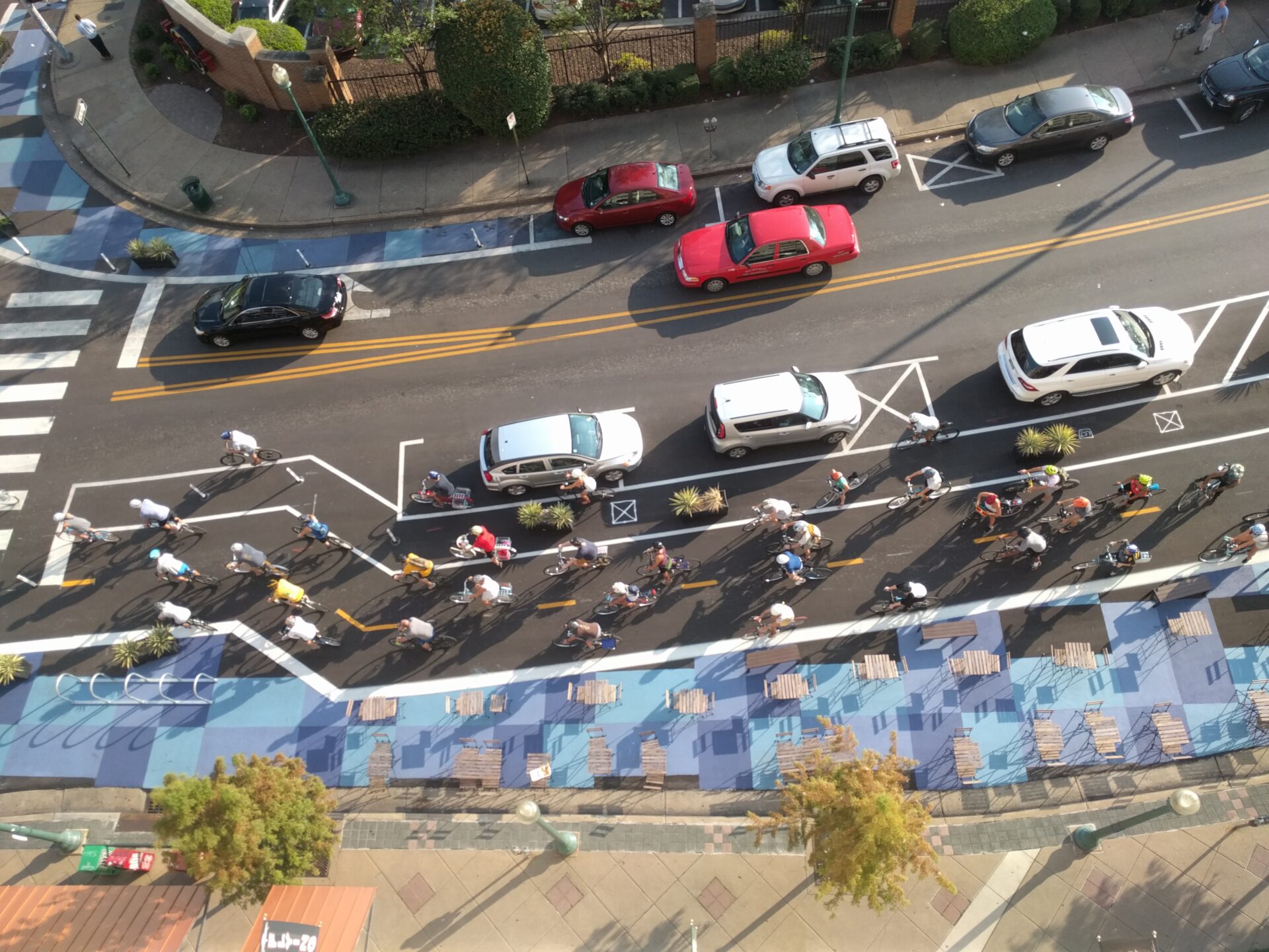 An aerial view of a street with an in-street plaza featuring public seating. People are biking in two-way parking protected bike lane. Cars in the travel lane approach an intersection.