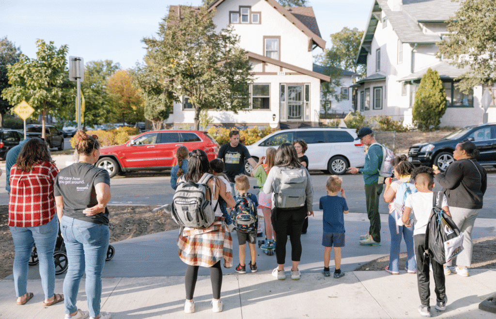 A group of adults and children gather on a sidewalk in a residential neighborhood.