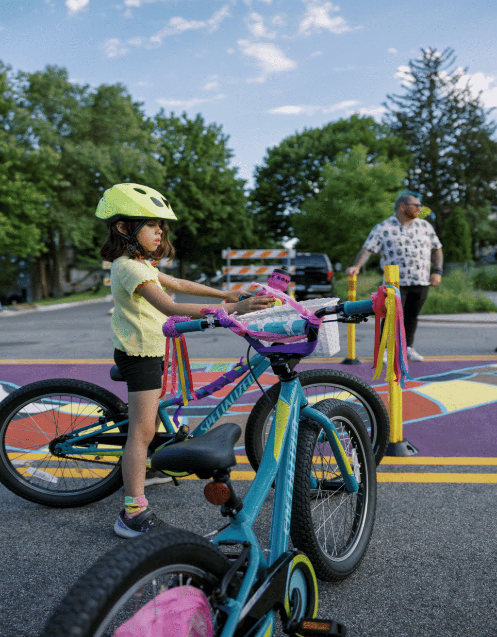 A young child in a yellow shirt wears a yellow helmet while standing over a teal balance bike.