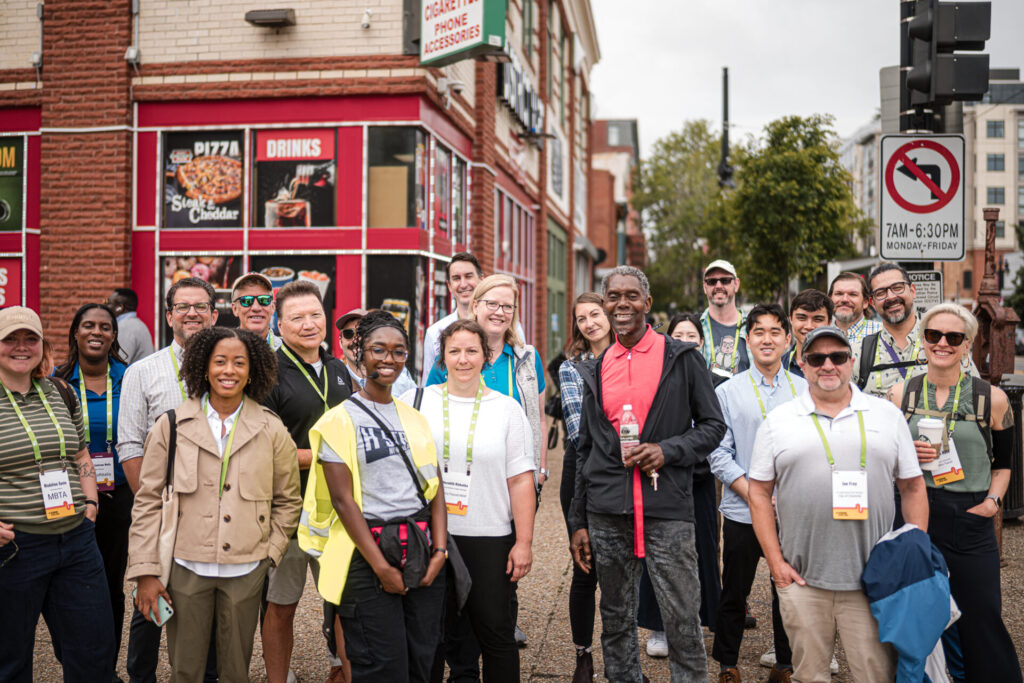 A few dozen attendees of Designing Cities 2025 in Washington DC pose together, smiling on a DC street
