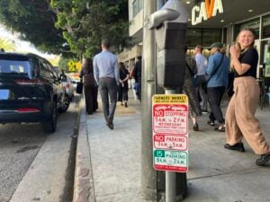 People walk on a sidewalk. The sign on the curb says "Farmers Market area special restrictions" along with parking and stopping regulations