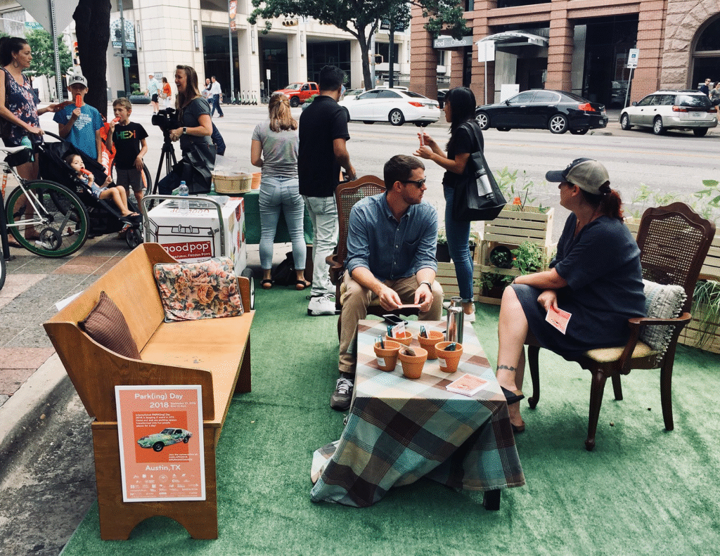 PARKing Day installation on Congress Ave in Austin, TX in 2018. A parking spot is covered in astroturf with a table and seating around. People are gathered in the space and chatting.