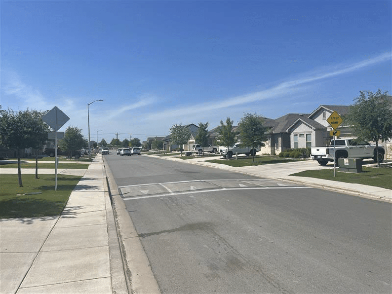 An asphalt speed hump is present on a residential street with concrete sidewalks and driveways.
