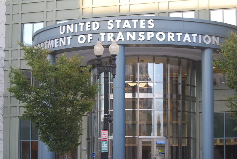 The entrance to the U.S, Department of Transportation in Washington, D.C. features a rounded glass portico with blue columns and a blue entablature. The department's name is prominently featured in silver letters.