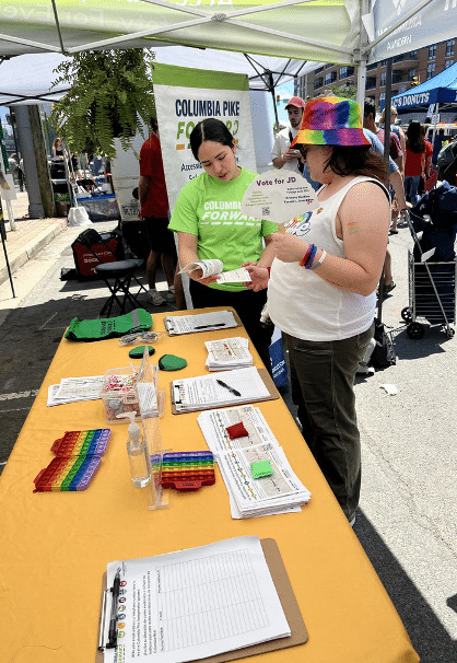 Two people are talking to each other in front of a table with a yellow tablecloth. They appear to be outside at a street fair. One of the people is wearing a lime green shirt with the words "Columbia Forward" on it.