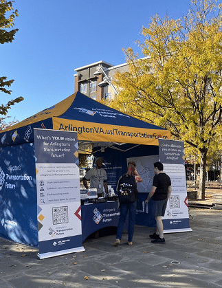 A blue and yellow tent with is on a plaza. Two pop-up signs flank the tent, with the words "What's YOUR vision for Arlington's transportation future?" at the top. Two people have approached the tent, and inside a man is standing behind a table.